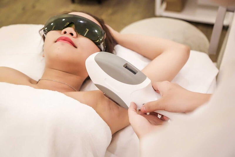 Người phụ nữ đeo kính bảo hộ, nằm thư giãn trong khi được triệt lông bằng thiết bị laser tại spa.