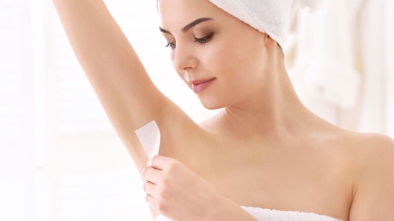 Người phụ nữ dùng miếng dán tẩy lông dưới cánh tay trong phòng tắm, đầu quấn khăn trắng.