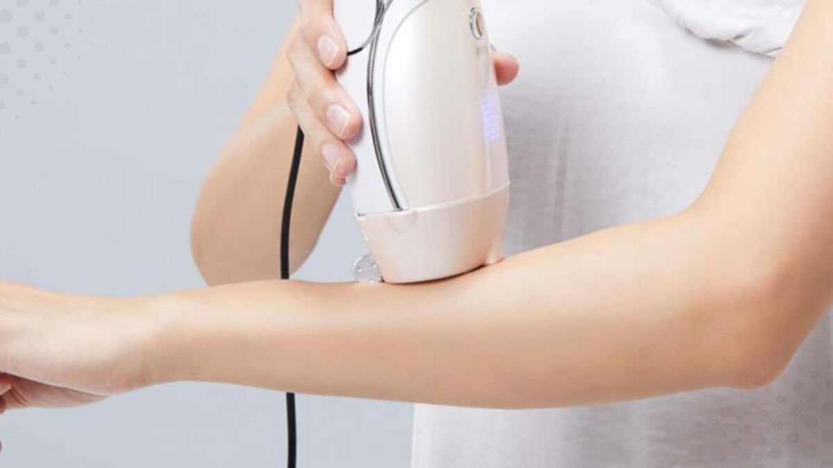 Người cầm thiết bị tẩy lông tay màu trắng, sử dụng trên cánh tay, nền màu xám.