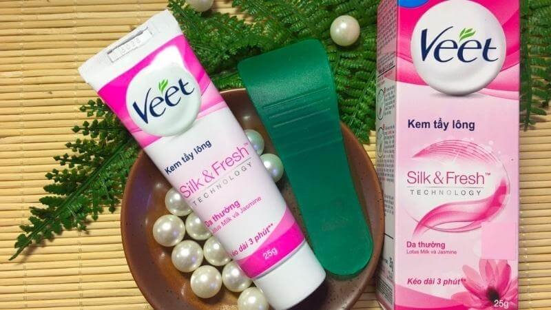 Hình ảnh một sản phẩm kem tẩy lông Veet, cùng với dụng cụ và hộp đựng, trên nền các viên ngọc trai và lá cây.