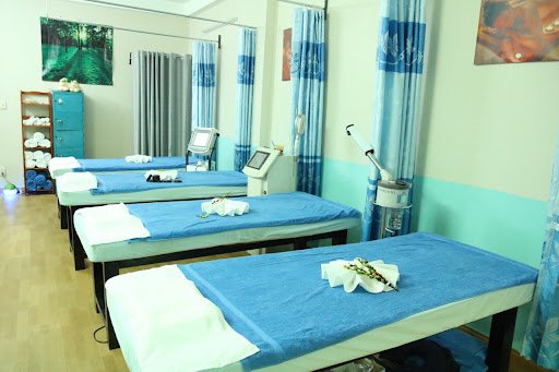 Phòng spa với bốn giường massage phủ khăn xanh, thiết bị chăm sóc da và rèm xanh trang trí.