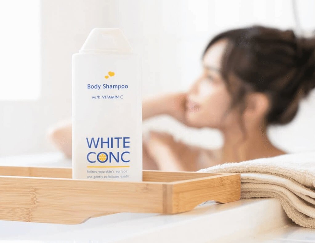 Chai sữa tắm White Conc trên kệ gỗ, nền mờ là một người đang tắm và khăn tắm xếp gọn.