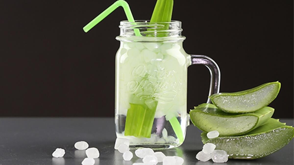 Cốc thủy tinh đựng nước nha đam với đá viên và ống hút, bên cạnh là các lát nha đam tươi trên nền đen.
