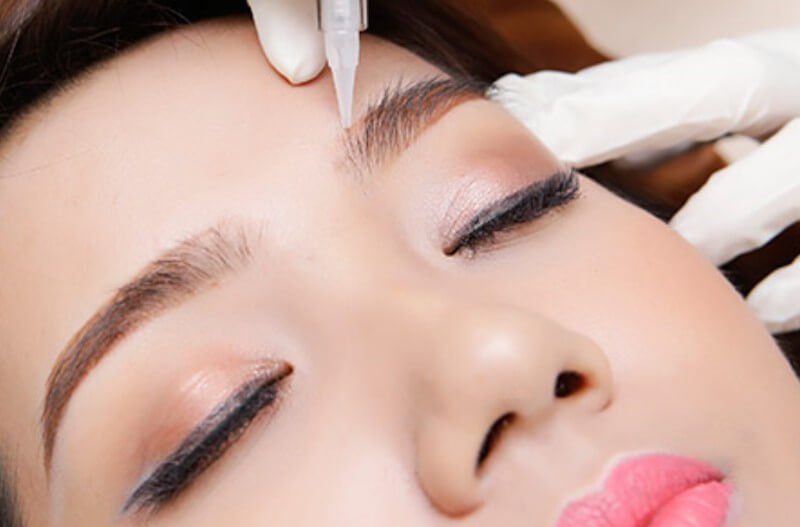 Người đang được thực hiện liệu trình chăm sóc lông mày với dụng cụ chuyên dụng trên trán, đeo găng tay trắng.