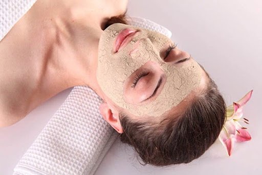 Người nằm thư giãn với mặt nạ đất sét trên mặt, đầu tựa trên khăn trắng, bên cạnh có hoa hồng.