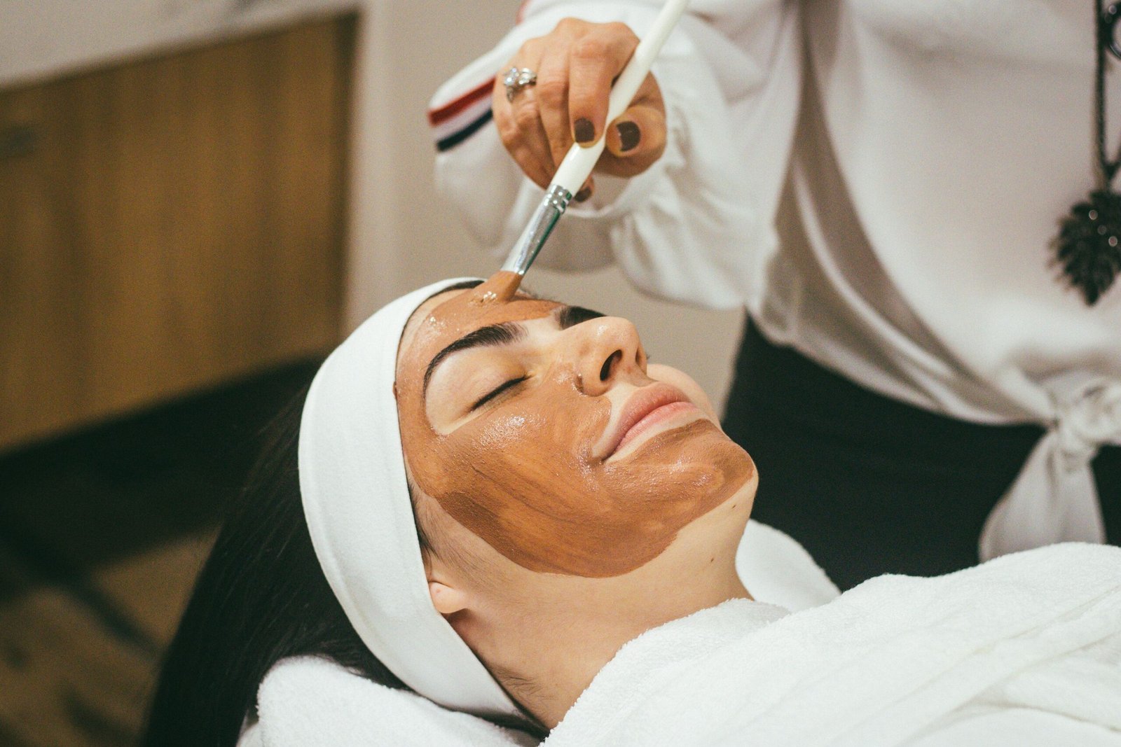 Người được đắp mặt nạ bùn trên mặt, nằm thư giãn trong spa, khăn trắng quấn quanh đầu.