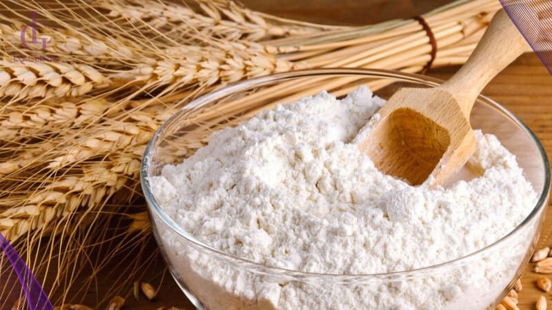 Bát bột mì với muỗng gỗ trên nền bó lúa mì khô.