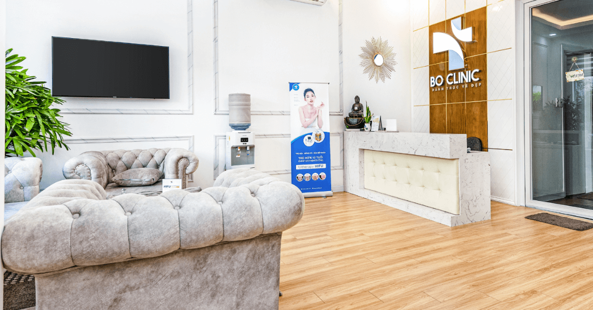 Phòng chờ sang trọng với ghế sofa, quầy lễ tân, máy nước và cây xanh, có biển hiệu và cửa kính.