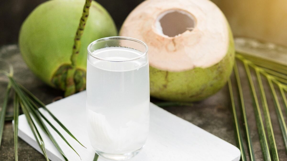 Ly nước dừa tươi đặt trên bàn cạnh hai quả dừa xanh và vài chiếc lá dừa.