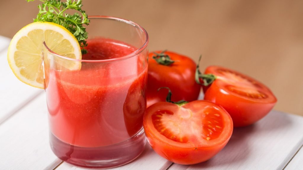 Ly nước ép cà chua trang trí lát chanh, bên cạnh là cà chua tươi trên bàn gỗ trắng.