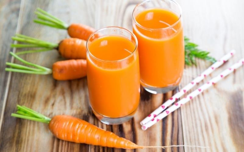 Hai ly nước ép cà rốt đặt trên bàn gỗ, bên cạnh là vài củ cà rốt tươi và ống hút.