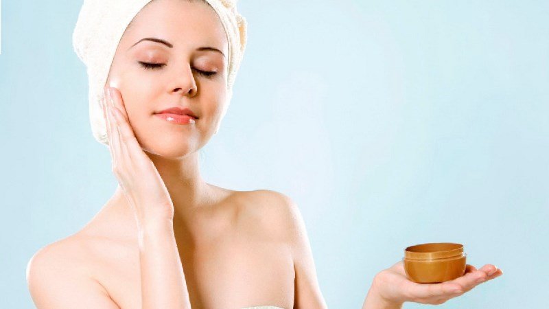Người phụ nữ quấn khăn, nhắm mắt thư giãn, tay cầm hộp kem dưỡng da, phông nền màu xanh nhạt.