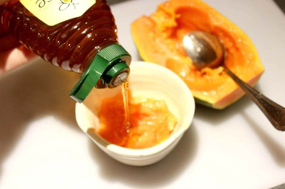 Đổ mật ong vào bát chứa đu đủ nghiền, bên cạnh là nửa quả đu đủ và một chiếc thìa.