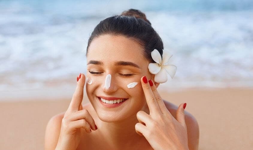 Người phụ nữ thoa kem chống nắng trên mặt, cười tươi, đứng trên bãi biển, hoa cài tóc.