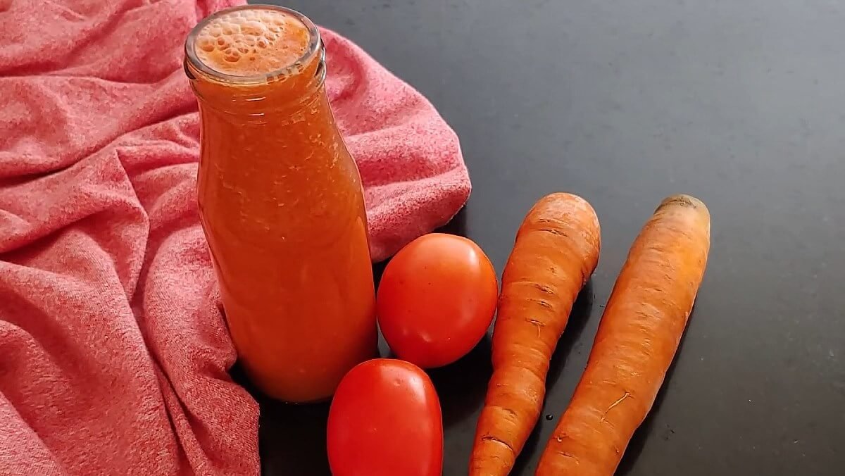 Chai nước màu cam cạnh hai quả cà chua và hai củ cà rốt trên nền vải đỏ.