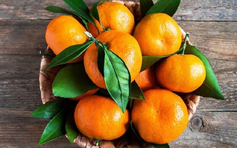 Rổ trái cam tươi, lá xanh, đặt trên nền gỗ nâu, tạo nên hình ảnh tươi sáng và tự nhiên.