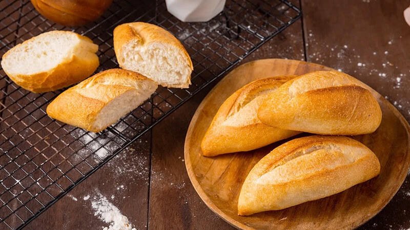 Ổ bánh mì vàng giòn trên đĩa gỗ và vỉ nướng, bên cạnh là một chút bột mì rắc trên bàn gỗ.