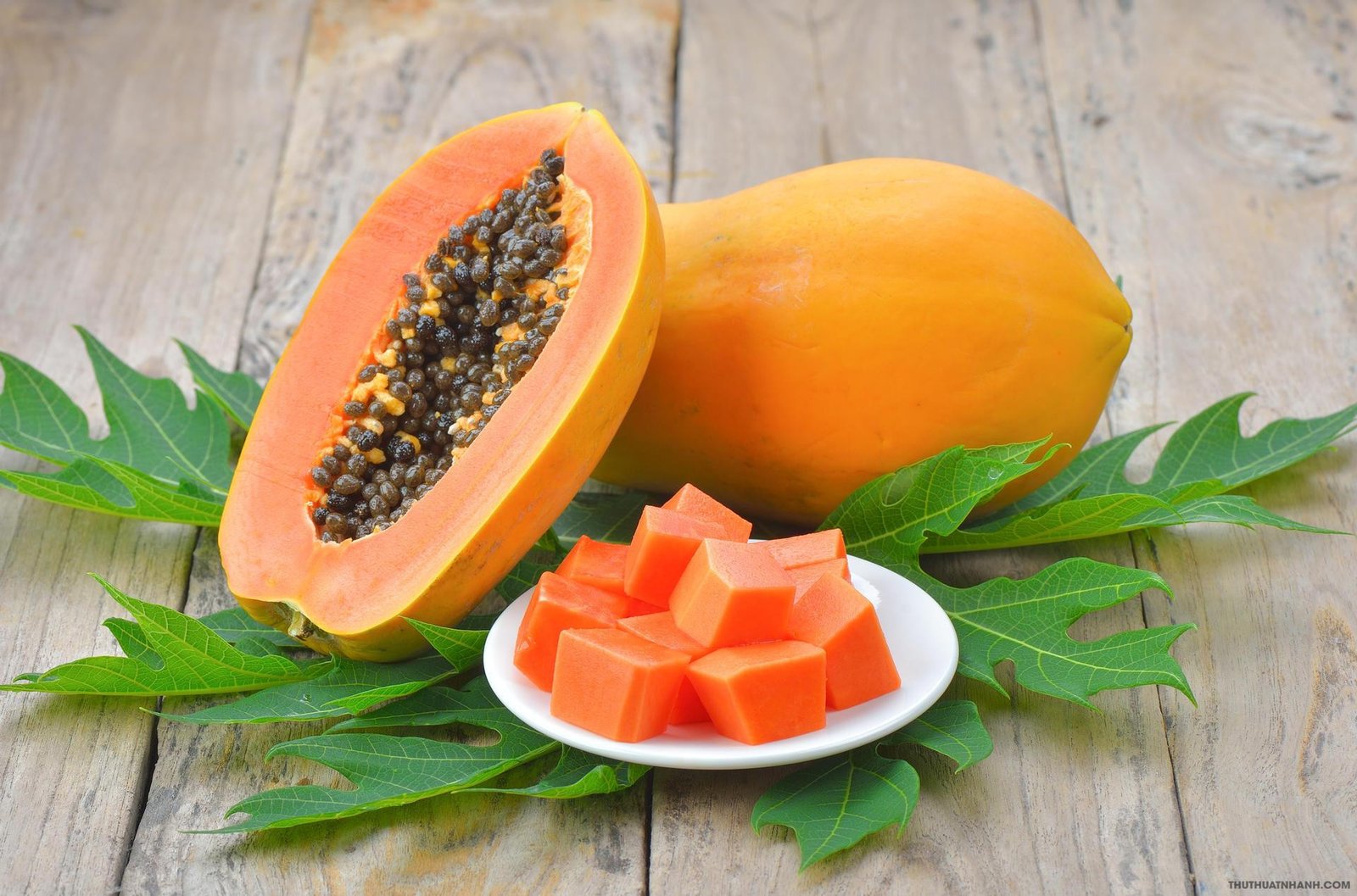 Đu đủ chín, một nửa cắt đôi để lộ hạt đen, đĩa đựng miếng đu đủ vuông, lá xanh trên bề mặt gỗ.