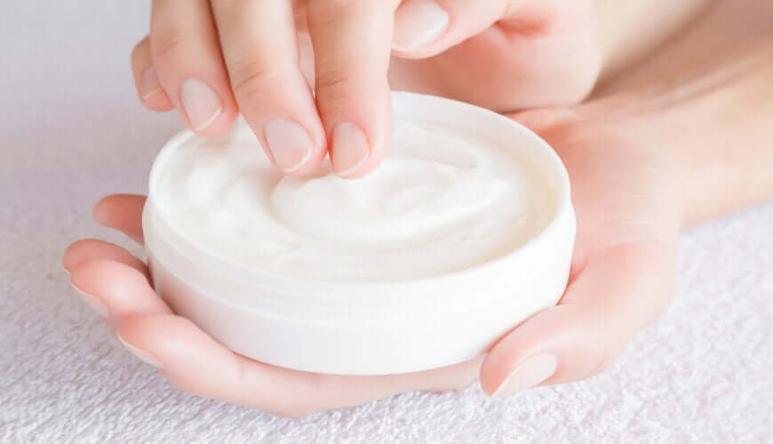Bàn tay đang lấy kem từ hộp trắng, đặt trên bề mặt màu sáng.
