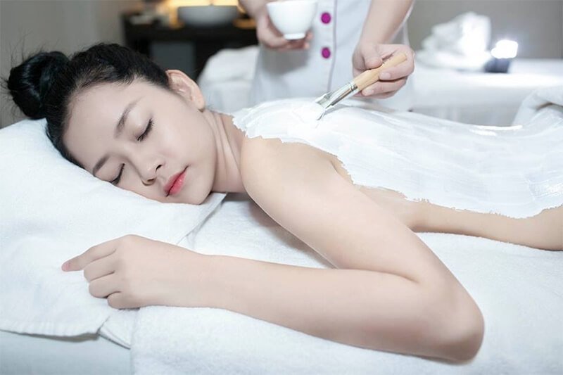 Người phụ nữ đang thư giãn trong spa, được chăm sóc da lưng với lớp kem trắng bằng cọ.