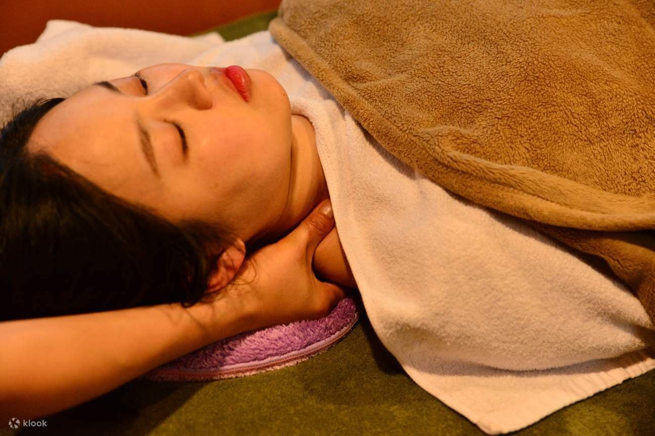 Người đang được massage cổ, nằm trên khăn mềm và đắp chăn, tạo cảm giác thư giãn.