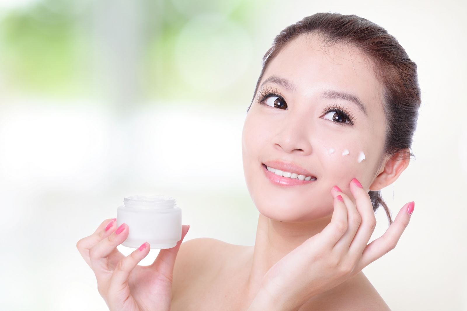 Người phụ nữ cầm hộp kem dưỡng, thoa kem lên má, nền phông mờ với tông màu sáng.