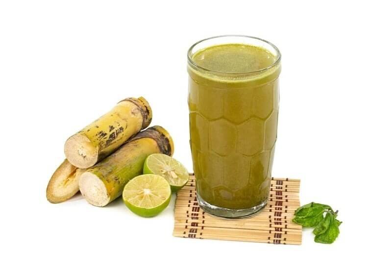 Ly nước mía tươi bên cạnh khúc mía và chanh cắt lát, trên nền trắng.