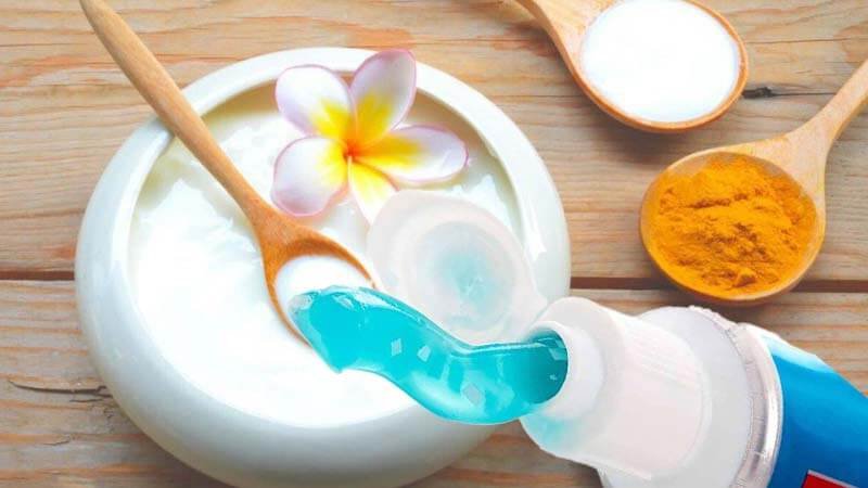 Bát sữa chua với hoa và thìa, bên cạnh là tuýp kem đánh răng và thìa bột nghệ trên nền gỗ.