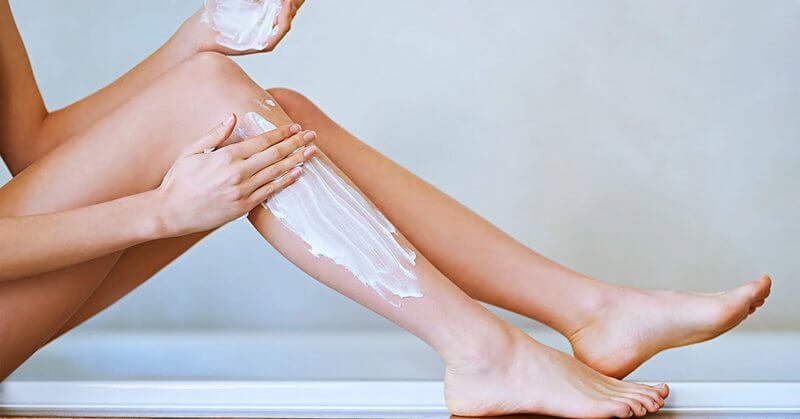 Người đang thoa kem dưỡng ẩm lên chân, ngồi trên bề mặt sáng màu.