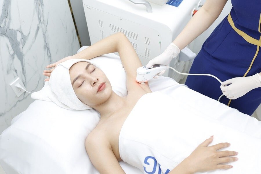 Người nằm trên giường spa, được chăm sóc vùng da dưới cánh tay bằng thiết bị thẩm mỹ, có khăn quấn đầu.
