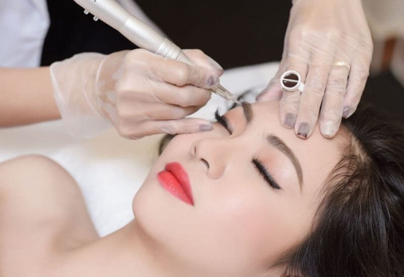 Người thợ đang thực hiện phun xăm thẩm mỹ lên chân mày cho khách hàng nằm thư giãn.
