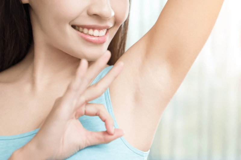 Người phụ nữ mỉm cười, giơ tay tạo dáng chữ "OK" cạnh vùng nách, mặc áo không tay màu xanh nhạt.