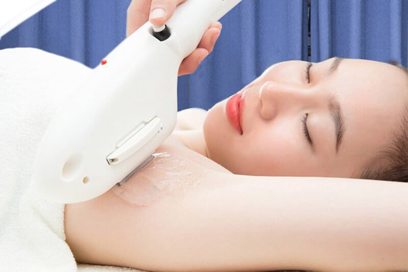 Người phụ nữ đang thư giãn trong khi triệt lông bằng máy laser ở vùng nách, phông nền màu xanh.