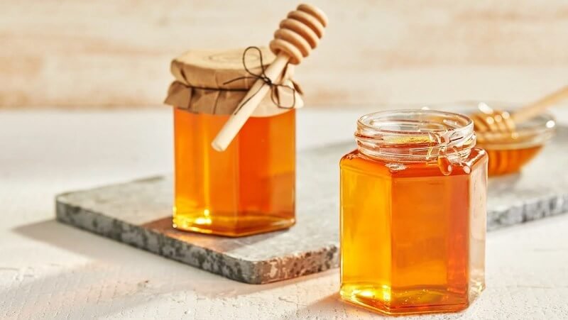 Hai lọ mật ong trên bàn, một lọ có nắp đậy và que khuấy, lọ kia mở nắp, ánh sáng chiếu qua tạo màu vàng óng.