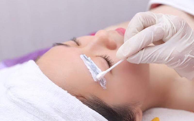 Người đang được thoa kem dưỡng lên lông mày bằng tăm bông, đội khăn trắng trên đầu.