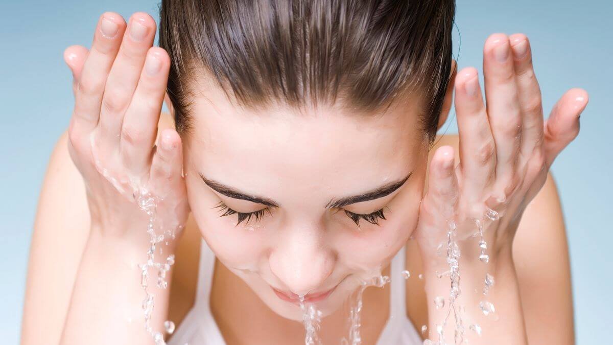 Người đang rửa mặt với nước, mắt nhắm, tay vốc nước lên mặt, nền xanh nhạt.