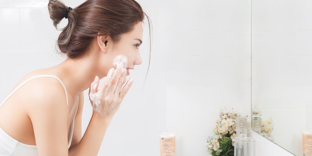 Người đang rửa mặt với bọt xà phòng trước gương trong phòng tắm sáng sủa, có hoa trang trí bên cạnh.