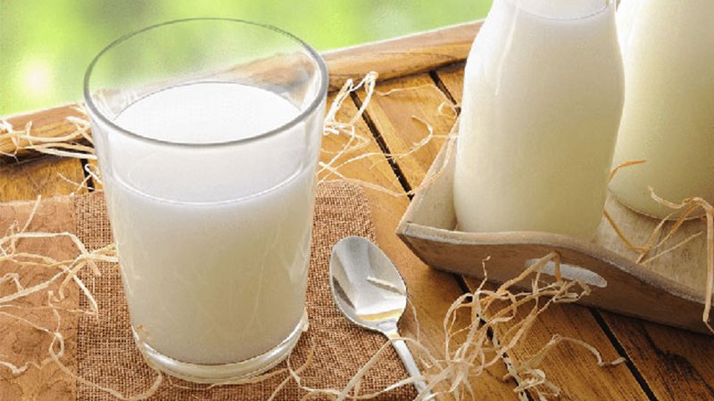 Ly sữa tươi đặt trên bàn gỗ cùng thìa kim loại và chai sữa, trang trí bằng rơm khô.
