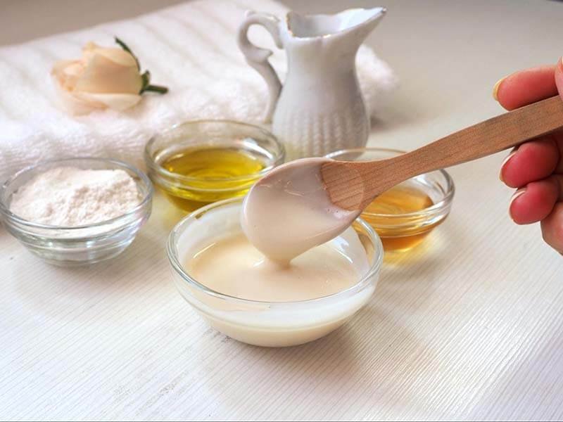 Người cầm thìa gỗ khuấy hỗn hợp trong bát, bên cạnh là các bát nhỏ chứa dầu, bột và mật ong.