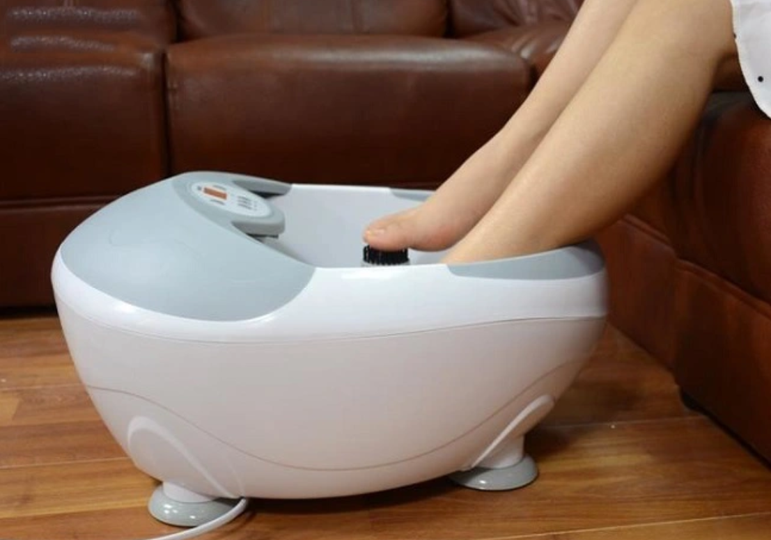 Bàn chân đang được ngâm trong máy massage chân màu trắng, đặt trên sàn gỗ cạnh ghế sofa.