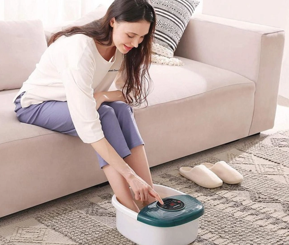 Người phụ nữ ngồi trên ghế sofa, ngâm chân trong bồn, tay điều chỉnh nút trên bồn, gần đó có đôi dép.