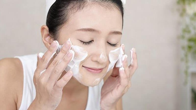Người phụ nữ đang rửa mặt bằng sữa rửa mặt tạo bọt, mắt nhắm và biểu cảm thoải mái.