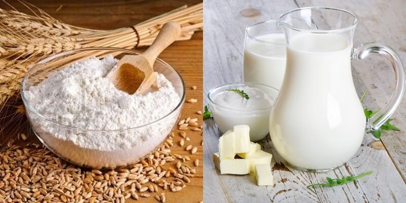 Bột mì trong bát cạnh lúa mì, bên cạnh là bình sữa, ly sữa, bơ và sữa chua trên bàn gỗ.