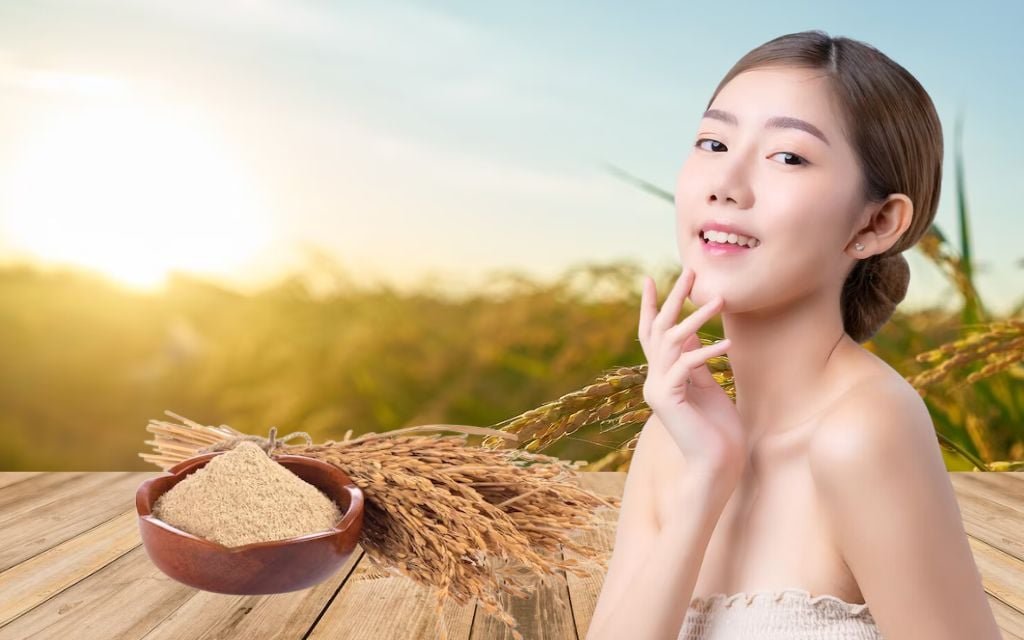Người phụ nữ cười nhẹ đứng cạnh bát bột và bó lúa trên bàn gỗ, nền là cánh đồng lúa và hoàng hôn.