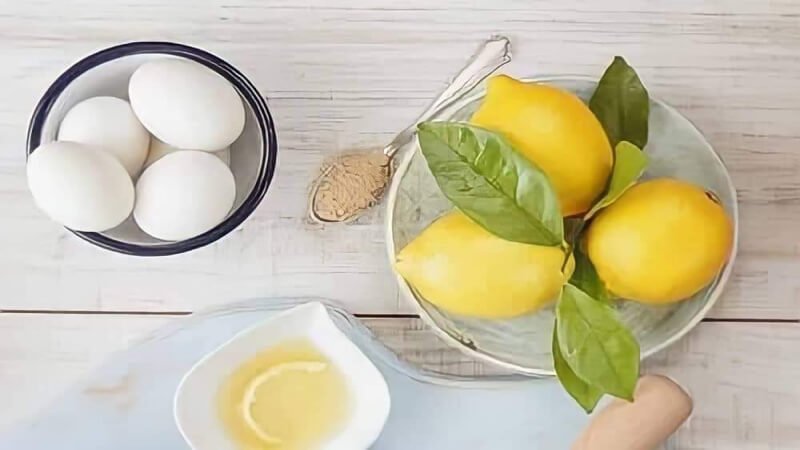 Bát trứng, đĩa chanh vàng có lá, muỗng bột, và bát nhỏ đựng chất lỏng trên bàn gỗ trắng.