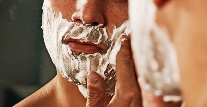 Người đang bôi kem cạo râu lên mặt, chuẩn bị cạo râu trước gương.