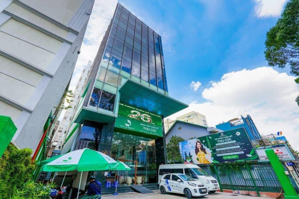 Tòa nhà kính cao tầng với biển hiệu xanh, trước cửa có ô tô và ô che nắng, trời xanh với mây trắng.