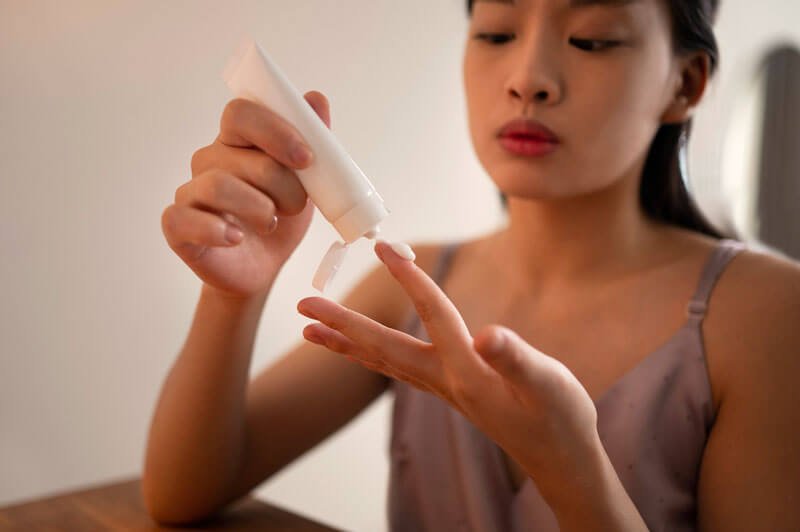 Người phụ nữ đang lấy kem từ một tuýp nhỏ lên ngón tay trong không gian nội thất ấm áp.