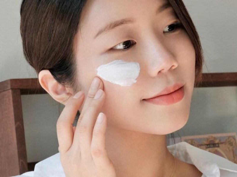 Người phụ nữ thoa kem dưỡng lên má, biểu cảm nhẹ nhàng, phông nền trong nhà.
