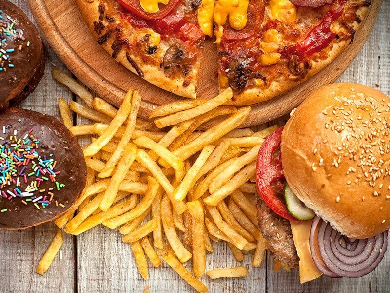 Pizza, bánh mì kẹp thịt, khoai tây chiên và bánh ngọt phủ sô cô la trang trí hạt màu trên bàn gỗ.